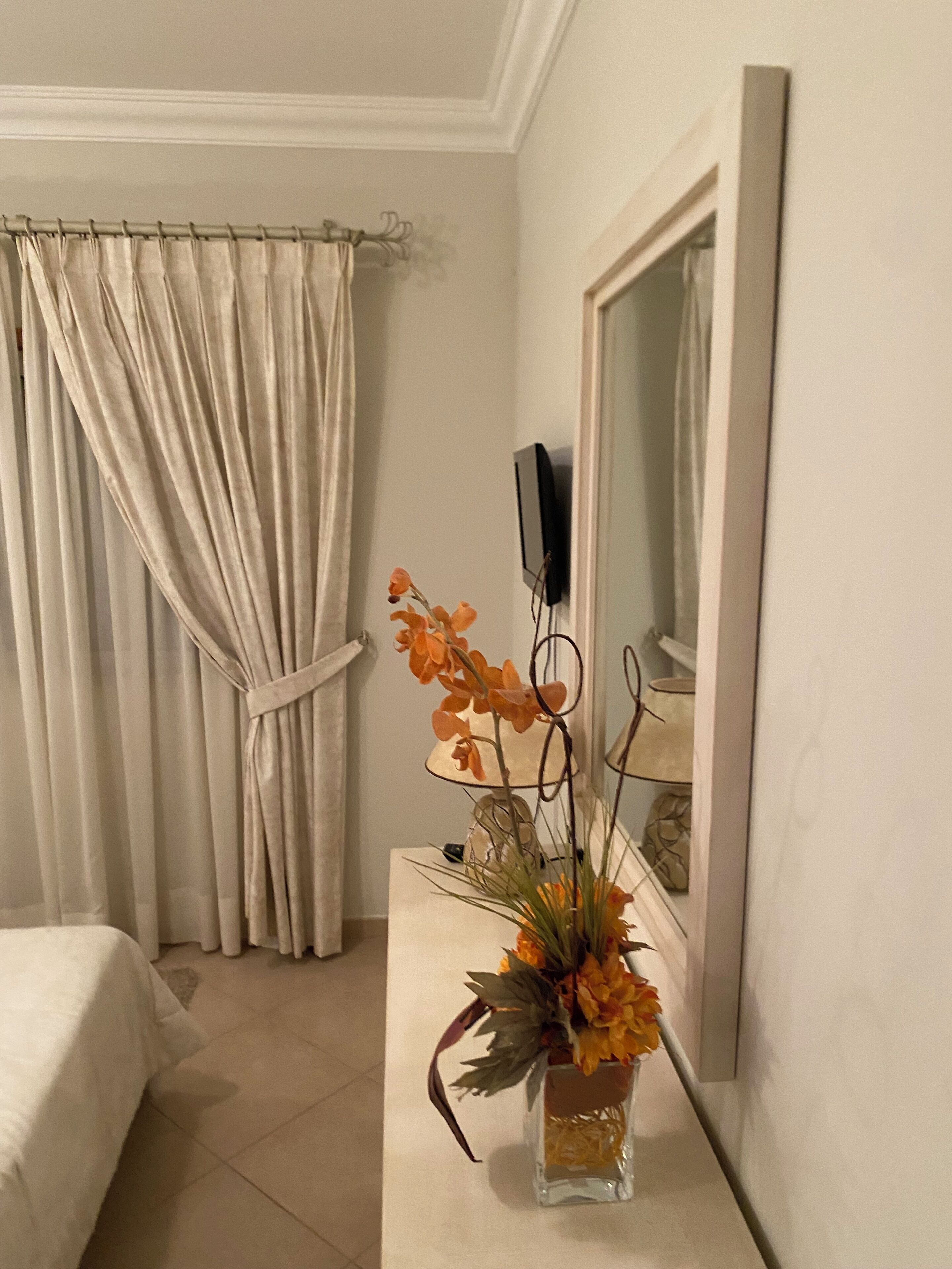A hotel room with a bed, a curtain, a vase with flowers, a mirror, and a television.