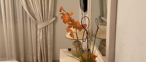 A hotel room with a bed, a curtain, a vase with flowers, a mirror, and a television.
