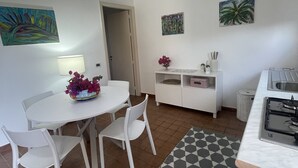 A modern kitchen with white cabinetry, a dining area with white chairs, and a kitchen island. There are two paintings on the walls, a white rug, and a tiled backsplash. - Casa al Mare, Villa Bella (Augusta)