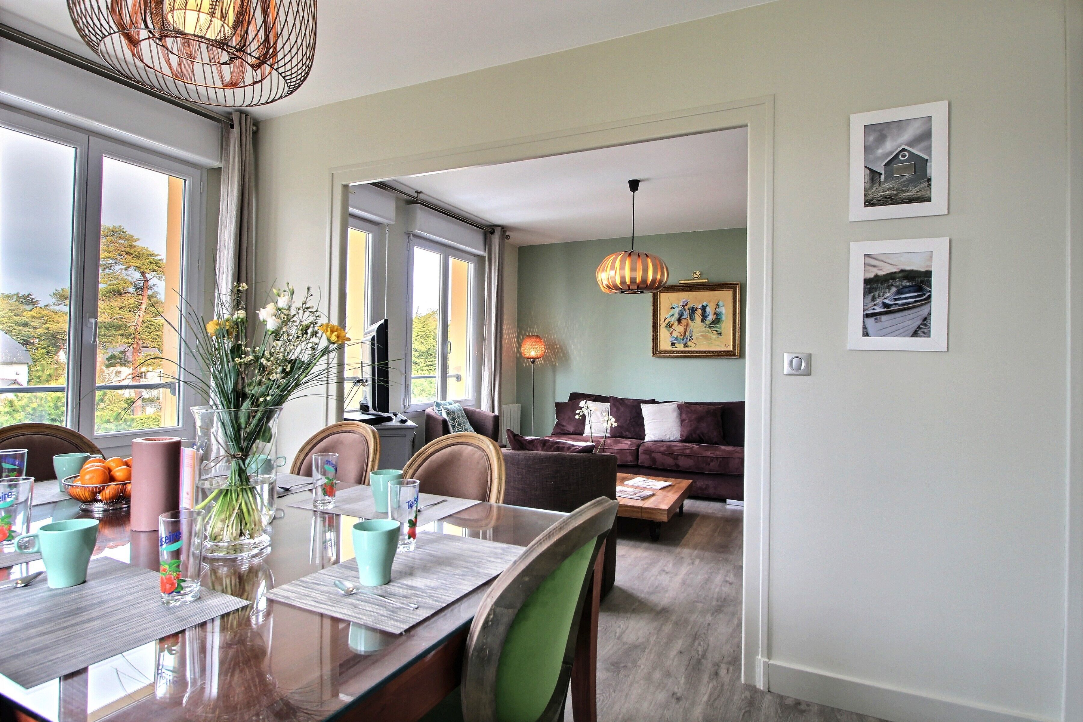 A dining area with a wooden table, chairs, and a vase of flowers. There is a sofa and a lamp in the background.