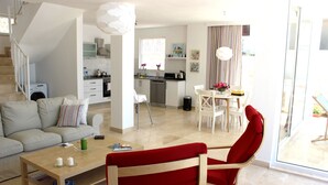 Un salon moderne avec un canapé, un fauteuil rouge, une table basse et un coin repas comprenant une table et des chaises. On aperçoit une cuisine avec une cuisinière et des placards au fond.