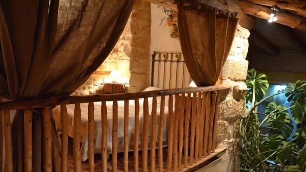 A rustic wooden balcony with a stone wall backdrop, a bed with a metal headboard, and a potted plant.