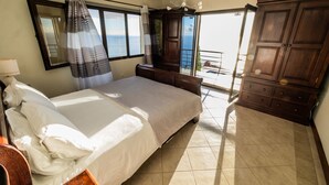 A bedroom with a bed, a wooden wardrobe, a window with curtains, and a view of the sea. - Secluded Sunset Villa on Cliff & 180° Ocean Views (Calheta)