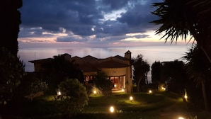 A house with a patio area, surrounded by trees and illuminated by multiple lights. - Secluded Sunset Villa on Cliff & 180° Ocean Views (Calheta)