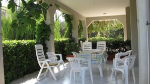 A covered patio with a table and chairs, surrounded by greenery and a view of the garden.
