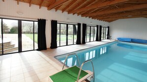 An indoor swimming pool with a wooden ceiling, large windows, and a blue pool edge.