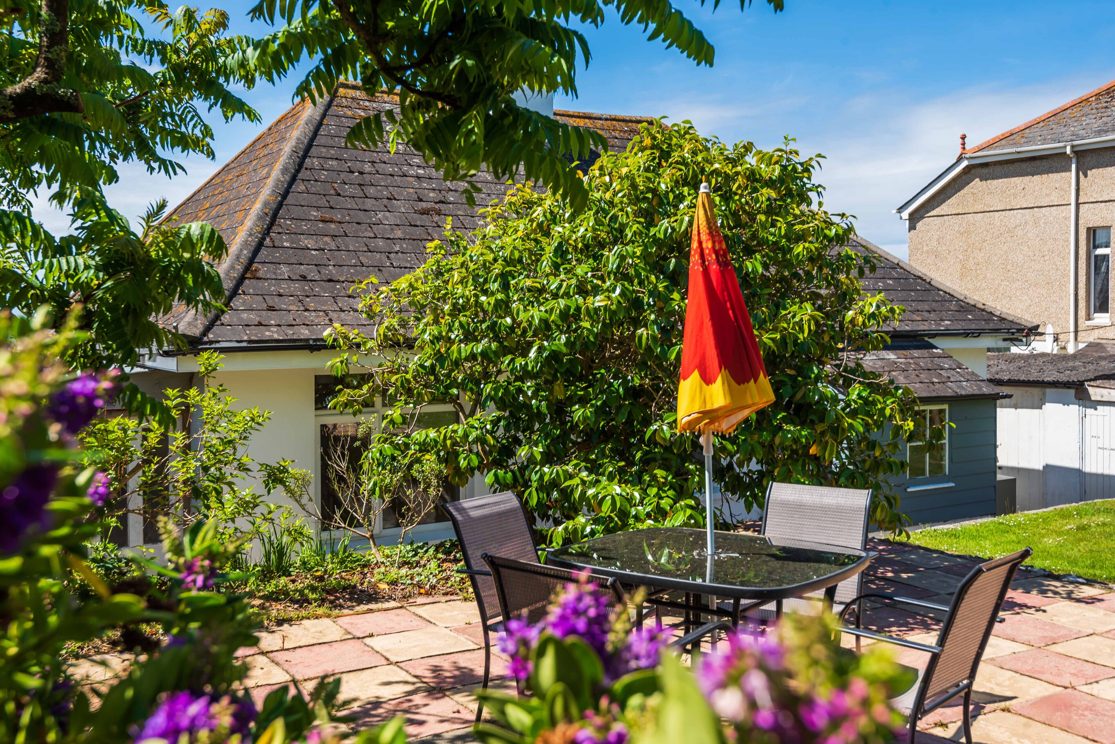 A patio with a table and chairs, surrounded by greenery and a house with a sloped roof.