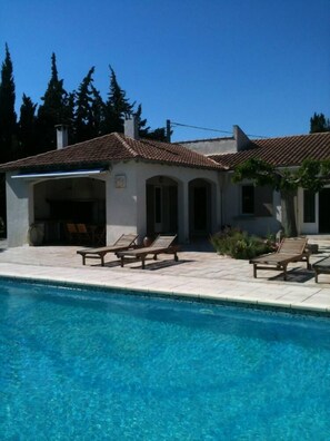 A poolside area with a clear blue pool, lounge chairs, and a building with a tiled roof.
