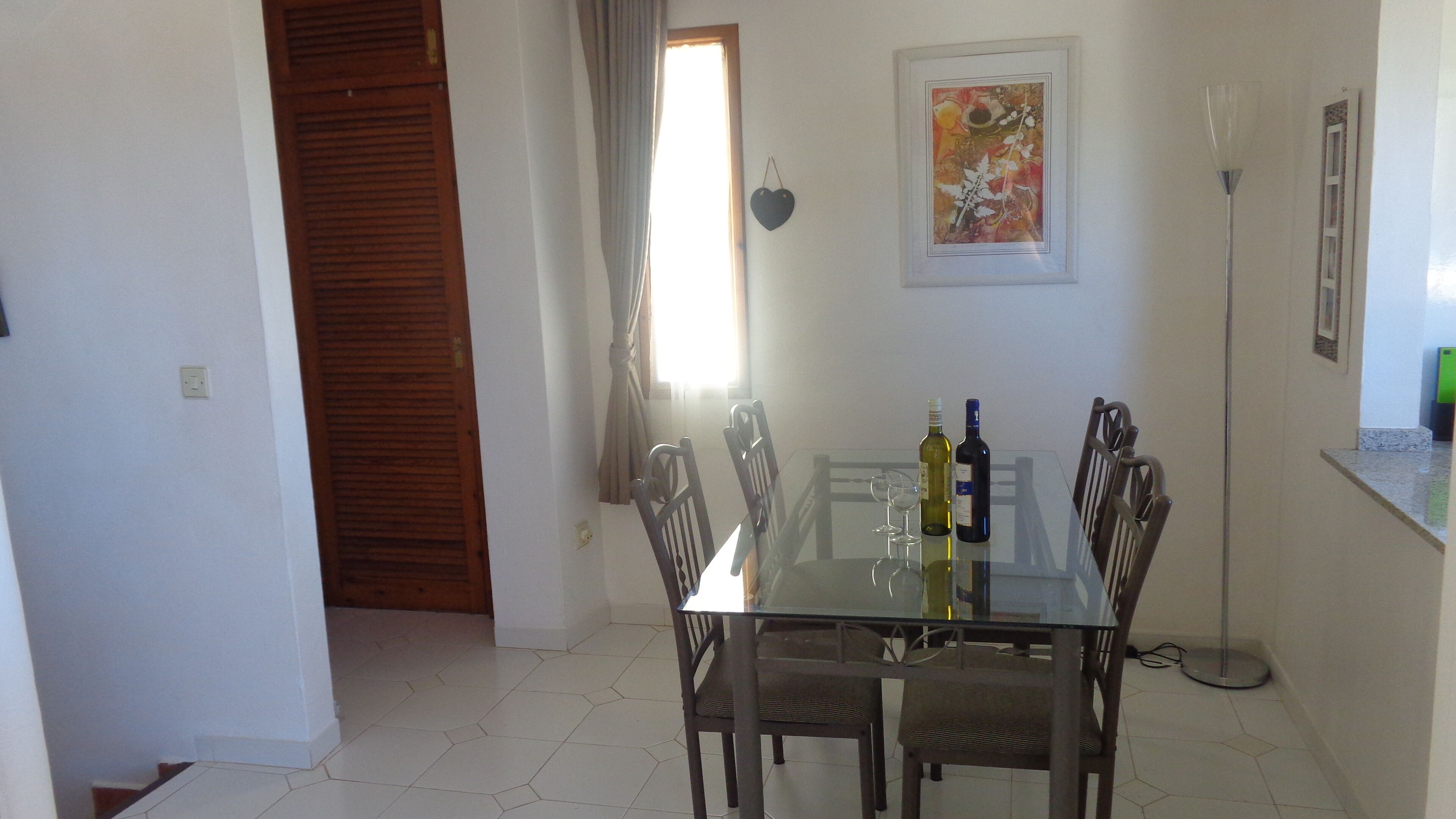 A dining area with a glass table and wooden chairs, two wine bottles, and a framed artwork on the wall.