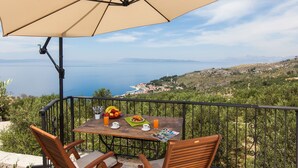 A wooden table with a beige umbrella, two wooden chairs, a plate of food, and a glass of orange juice on a balcony with a scenic view of the sea and hills. - Villa Oliva - exclusive gateway, luxurious serenity, stunning views, heated pool (Podgora)
