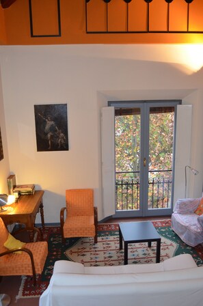 Una habitación con una mesa de madera, dos sillas, una mesita, un sofá, un balcón con barandilla y una puerta con paneles de vidrio.