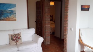 Een kamer met een witte bank, een decoratief kussen, een houten stoel en een deur naar een andere kamer.