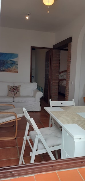 A living room with a white sofa, a coffee table, and a dining table with chairs. There is a painting on the wall and a door leading to another room.