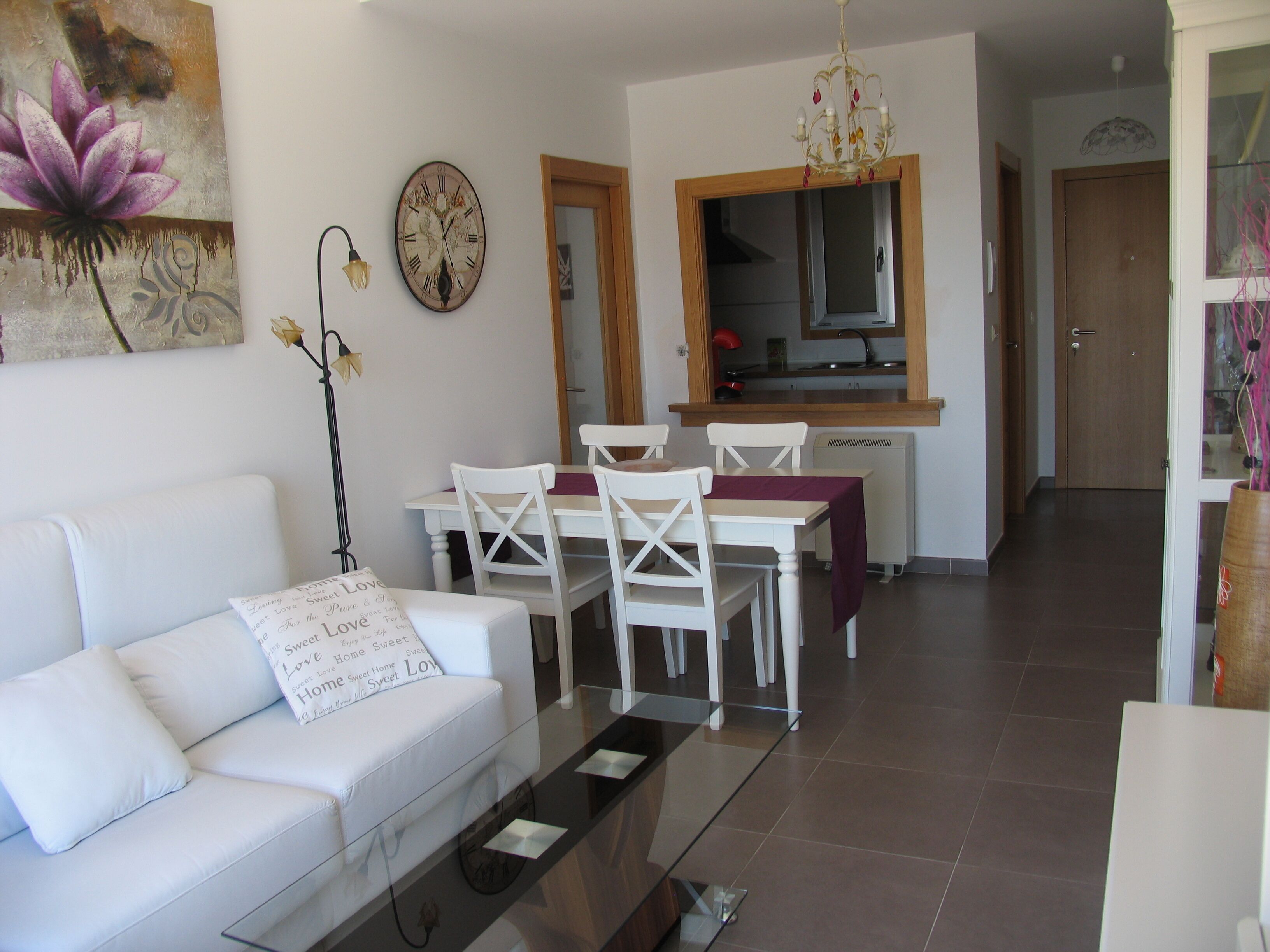 A modern living room with a white sofa, a dining table with chairs, a kitchen area, and a large mirror.