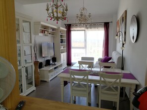 Una sala de estar moderna con televisor, mobiliario blanco, una araña y un comedor con mesa y sillas.