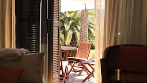 Ein Hotelzimmer mit einem Bett, einem Tisch mit Flasche und Gläsern sowie Blick auf Palmen durch eine offene Tür.