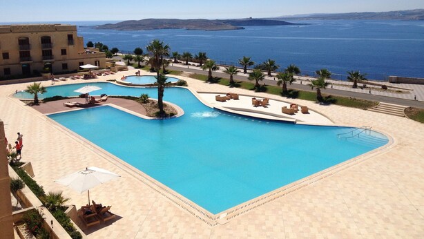 En pool på ett semesterhotell med flera avdelningar, palmer och utsikt över havet.