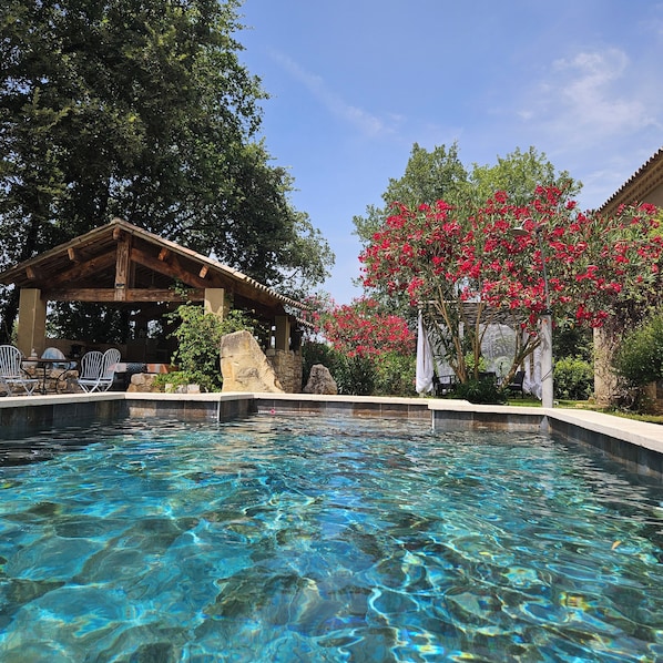 A clear swimming pool with a wooden pergola and chairs, surrounded by lush greenery and red flowering plants.