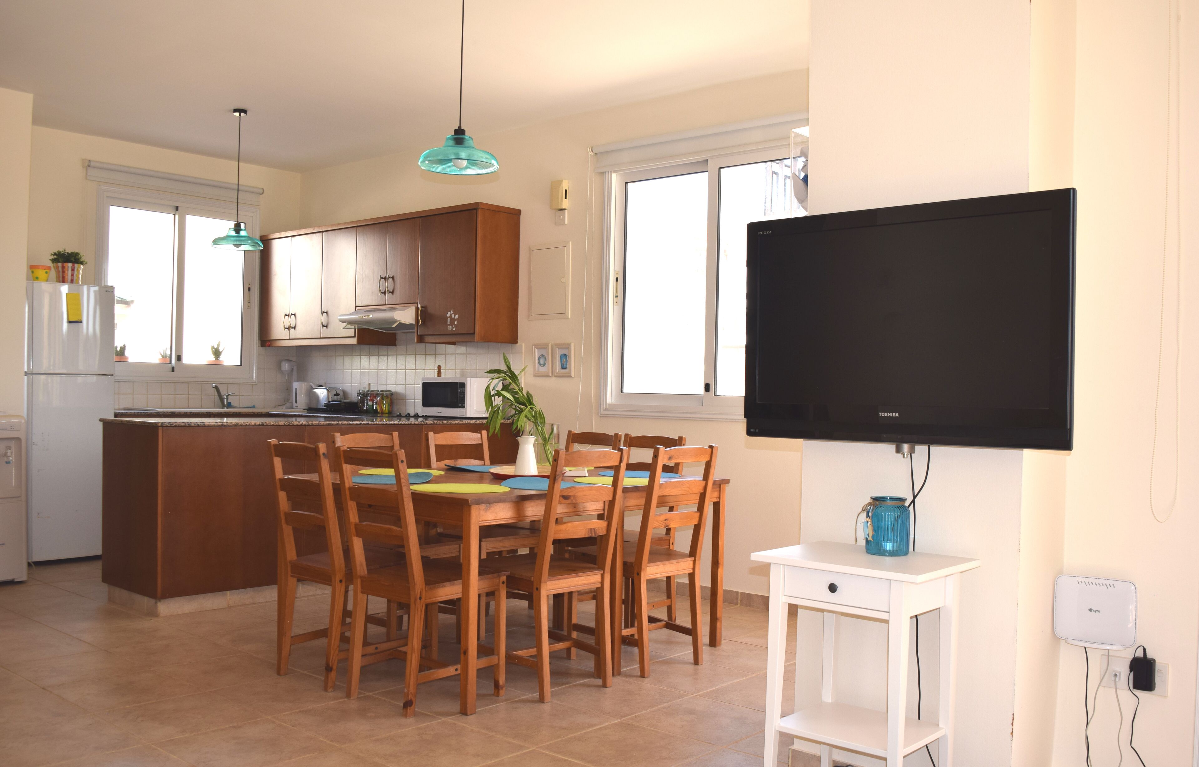 A modern kitchen with wooden cabinets, a dining table with chairs, a large flat-screen TV, and a small white cabinet.