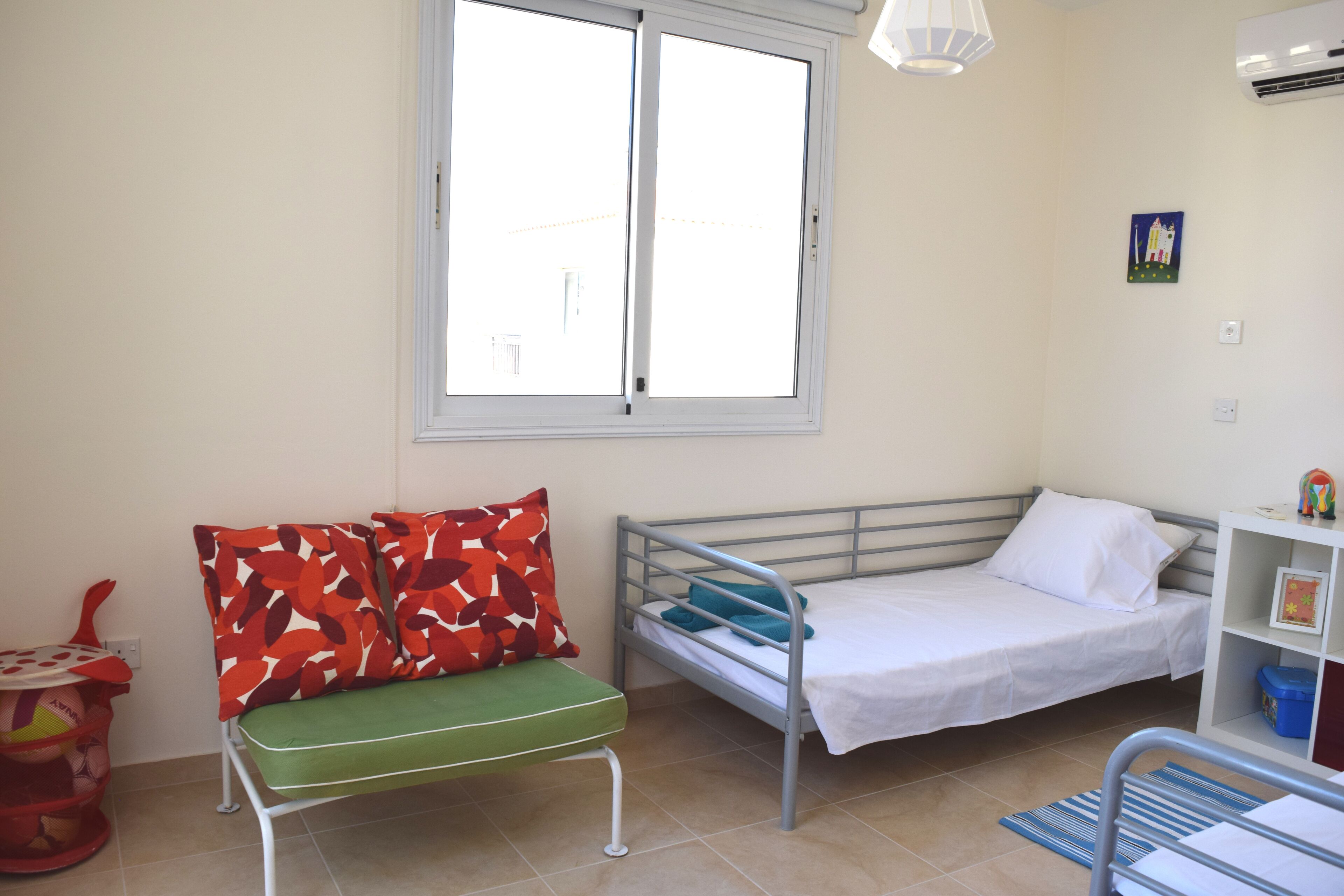 A small, well-lit room with a bed, a green cushioned bench, a window, and a shelf with a picture frame.