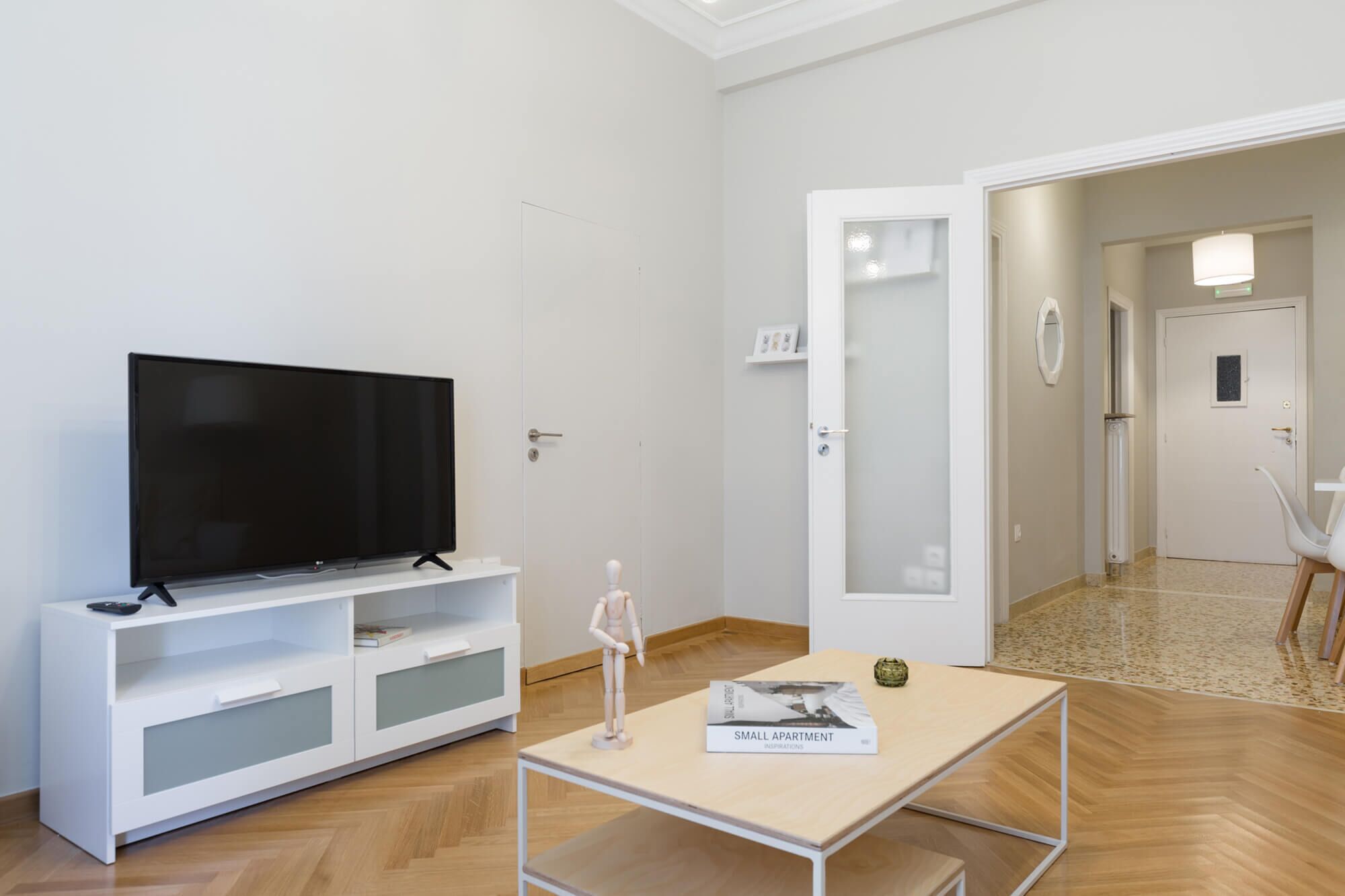 Ein modernes Wohnzimmer mit einem Flachbildfernseher auf einer weißen Kommode, einem kleinen Tisch mit einem Buch und einer Tür zu einem anderen Raum.
