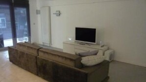 Una sala moderna con un sofá seccional marrón oscuro, un televisor de pantalla plana sobre un pedestal blanco y un ventanal que da al exterior.