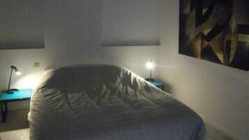 A bedroom with a bed covered in a grey blanket, a desk with a lamp, and a painting on the wall.