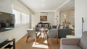 A modern living room with a flat-screen TV, grey sofas, a dining table, and a ceiling-mounted air conditioning unit.