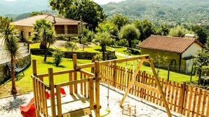 Un parque infantil con un conjunto de columpios y un tobogán de madera, rodeado por una cerca de madera, y una casa y abundante vegetación al fondo.