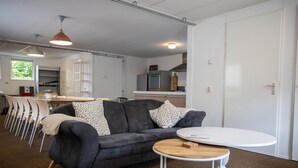 A modern living room with a grey sofa, a round wooden coffee table, and a dining area with white chairs and a table. - Spacious & cozy holiday bungalow "Frank" in wooded area (12 people) (Den Ham)