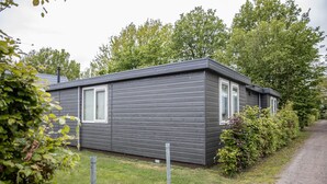 A single-story, grey wooden cabin with a large window on the front, surrounded by greenery and a gravel path. - Spacious & cozy holiday bungalow "Frank" in wooded area (12 people) (Den Ham)