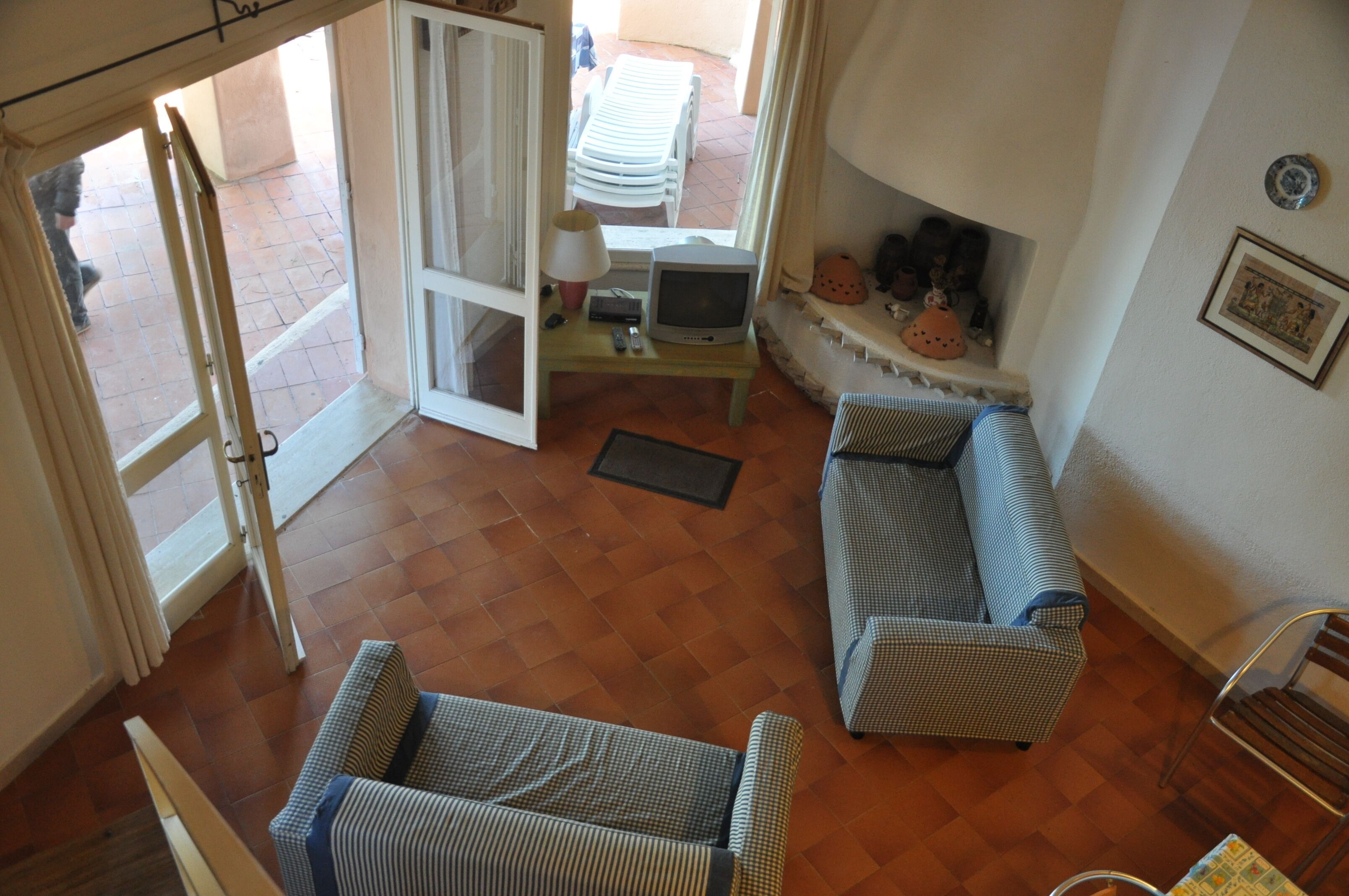 A living room with a sofa, a television, and a small table. There is a door leading to an outdoor area with a chair and a table.