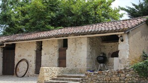 Un edificio rústico de piedra con techo de tejas, puerta de madera y una parrilla en el exterior.