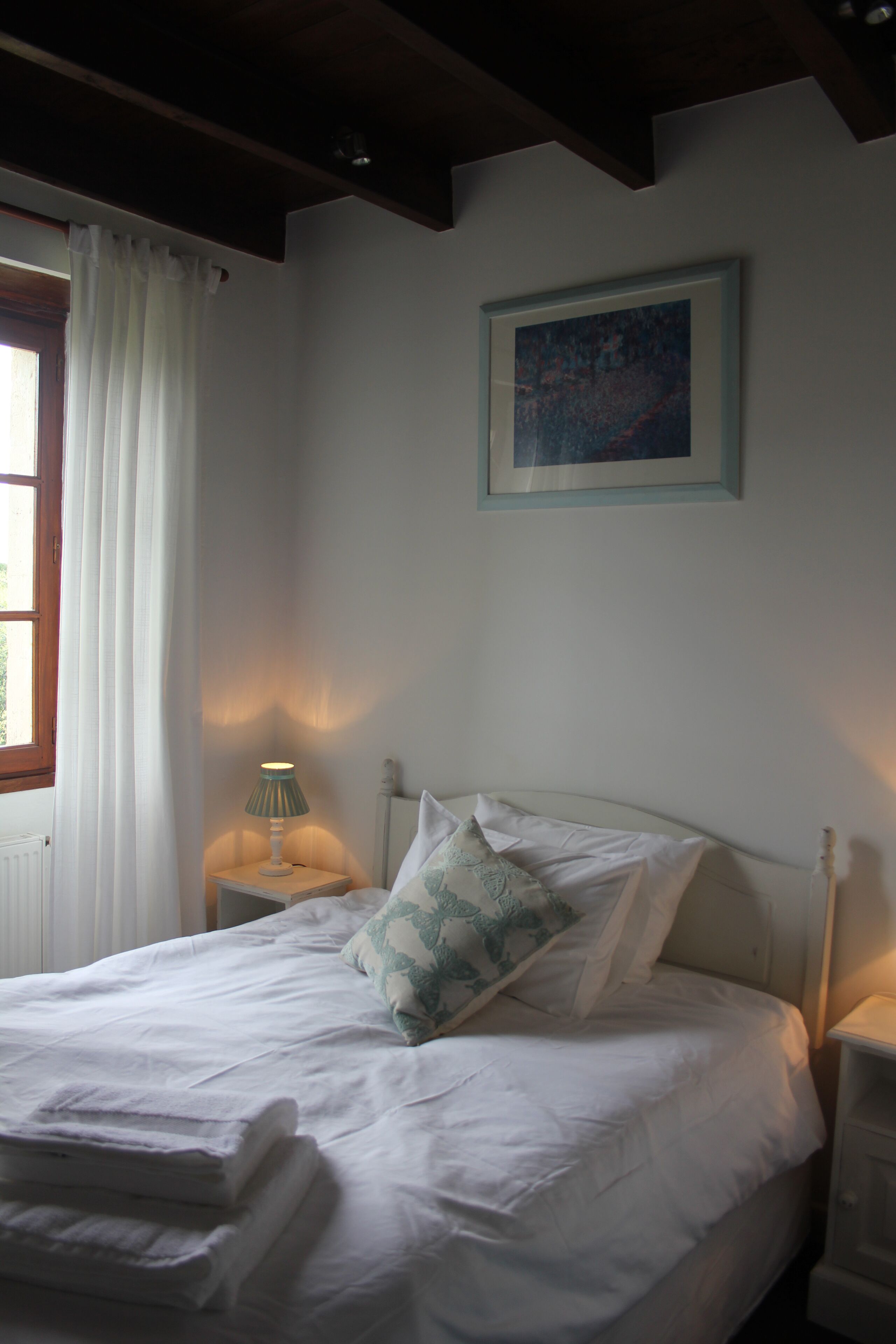 A neatly made bed with white linens and a folded towel at the foot. There is a bedside table with a lamp, a framed picture on the wall, and a window with white curtains.