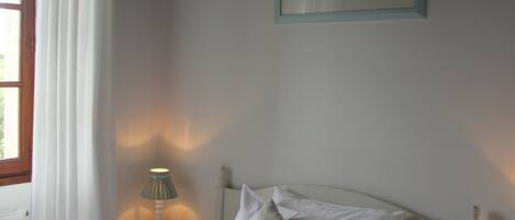 A neatly made bed with white linens and a folded towel at the foot. There is a bedside table with a lamp, a framed picture on the wall, and a window with white curtains.