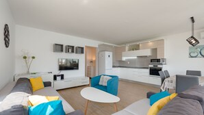 Una sala de estar moderna con un sofá, una mesita y un televisor. Se ve una cocina al fondo con un microondas y armarios.