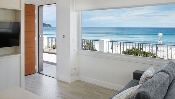 Una sala de estar con un ventanal que da a la playa, un televisor de pantalla plana en la pared y una puerta de madera que conduce a un espacio al aire libre.