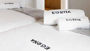 Una habitación de hotel con una cama, dos toallas con el nombre de marca 'EGONA', una puerta de madera que da a un baño con lavabo y espejo.