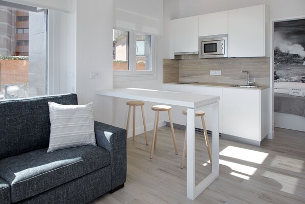 A modern kitchen with white cabinets, a wooden countertop, and a built-in microwave. There is a grey sofa with a white pillow and wooden stools. - Mollarri apartment by Egona (Zarautz)