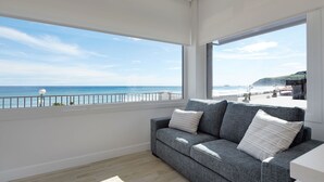Una sala de estar moderna con un sofá gris, piso de madera y un amplio ventanal con vista a la playa y al océano.