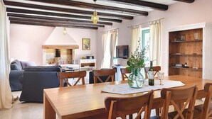 Uma sala de estar acolhedora com lareira, vigas de madeira, uma mesa de jantar com cadeiras e uma televisão ao fundo.