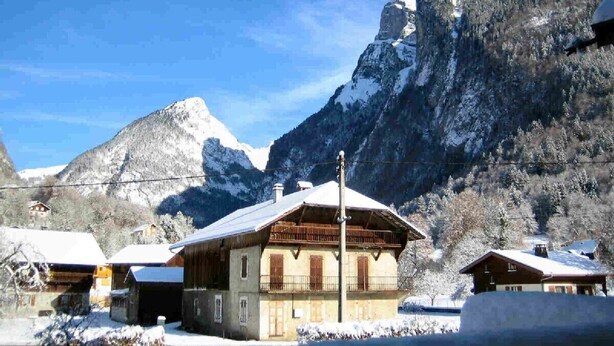 Un refugio de montaña con fachada de madera, tejados nevados y montañas cubiertas de nieve al fondo.