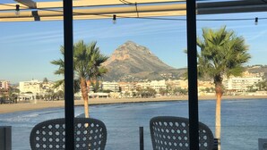 A view from a covered outdoor seating area with two chairs and a table, overlooking a beach with palm trees and a mountain in the background. - Detached Villa Close to Arenal Beach, Javea -Stunning, Very Private Villa & Pool (El Tosalet)