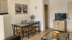Un coin repas avec une table et des chaises en verre, un ventilateur et un téléviseur posé sur un meuble.