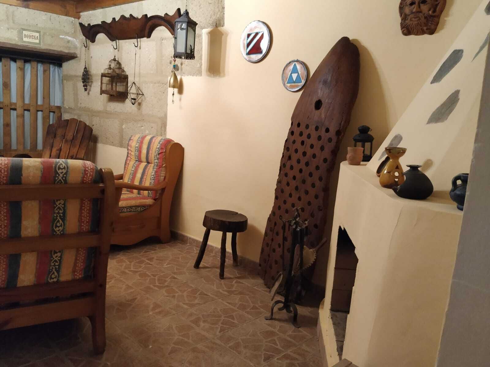 Una habitación acogedora con una silla de madera, un cojín estampado, un taburete pequeño y una tabla de surf apoyada en la pared.