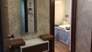 A hotel room with a wooden vanity, a mirror, a bedside table, a bed with a grey duvet, and a nightstand with a lamp. - I Love Alegria - cosy Studio (Porto)