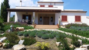 A two-story house with a covered porch, a garden with various plants and flowers, and a clear blue sky. - Cozy, quiet house with Pool, Wifi and sea view (Son Servera)