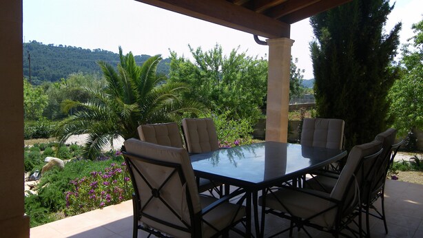 An outdoor patio with a table and chairs, surrounded by greenery and a view of hills. - Cozy, quiet house with Pool, Wifi and sea view (Son Servera)
