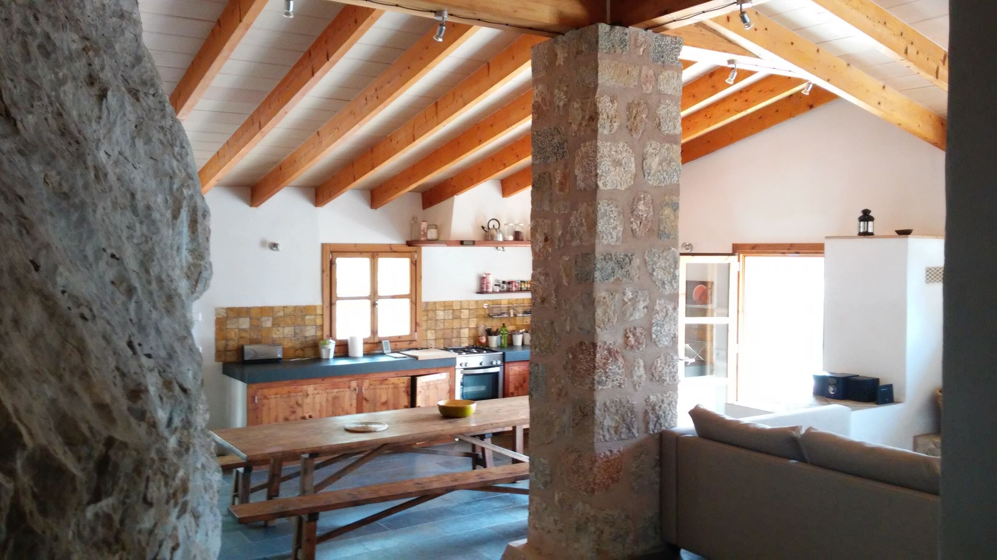 A rustic kitchen with exposed wooden beams, a stone wall, and a stone floor.