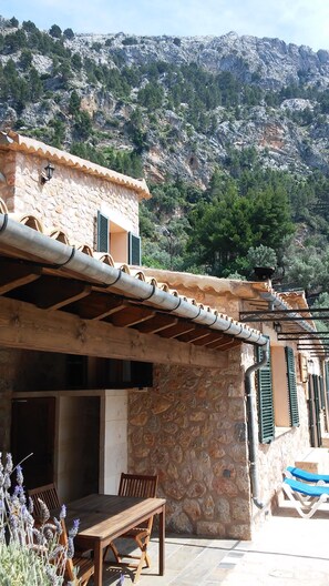 Un edificio de piedra con una pérgola de madera, asientos al aire libre y montañas al fondo.